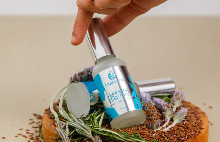 Hand reaching for a Müllwood Hydrating Hair Mist bottle on a board with flaxseed, rosemary and lavender